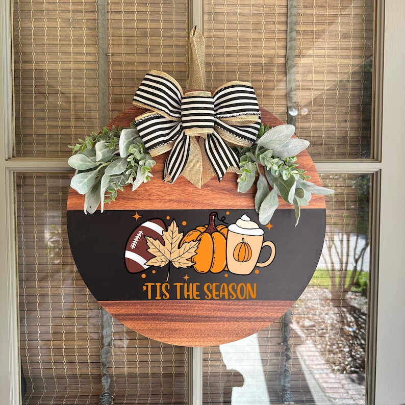 May include: A round wooden fall-themed wreath with a black and brown striped bow and greenery. The wreath features a football, pumpkins, a leaf, and a pumpkin spice latte graphic. The text "'Tis the Season" is printed on the bottom.