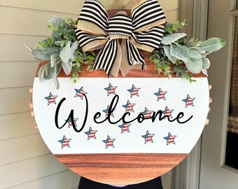 May include: Round wooden welcome sign with a white and wood grain design. The sign features the word "Welcome" in black script, surrounded by stars with red, white, and blue stripes. A large bow with black and white stripes and greenery accents the top.