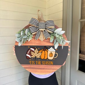 May include: Round wooden fall-themed door hanger with a black and brown design. It features a football, pumpkins, and a coffee cup graphic. The text reads "'Tis the Season". The hanger is decorated with a striped bow and greenery.