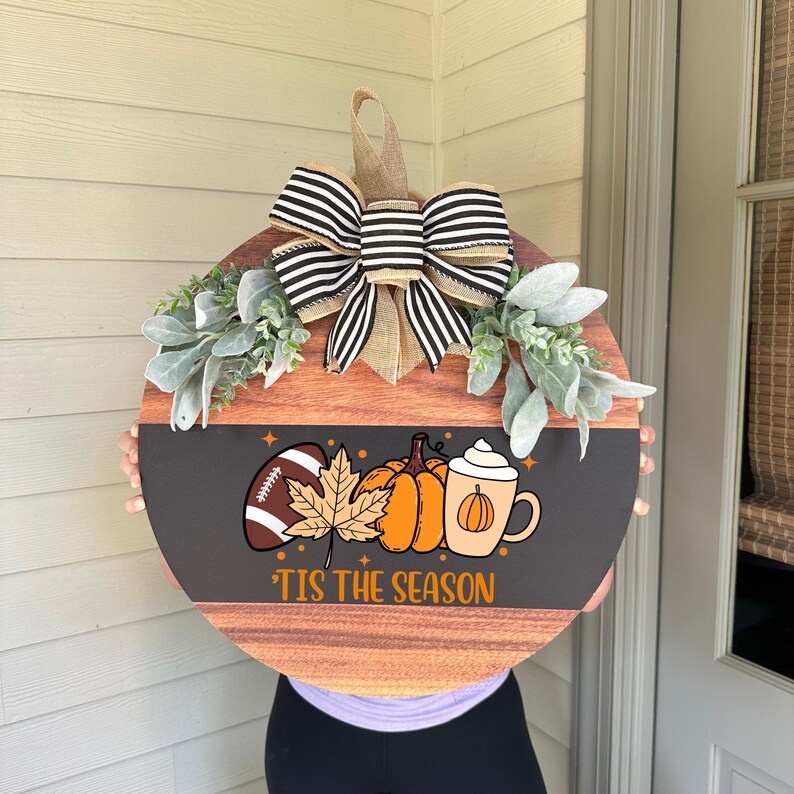 May include: Round wooden fall-themed door hanger with a black and brown design. It features a football, pumpkins, and a coffee cup graphic. The text reads "'Tis the Season". The hanger is decorated with a striped bow and greenery.