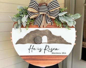 May include: A round wooden Easter door hanger with a white and brown design. The design includes a depiction of the empty tomb, the text "He is Risen Matthew 28:6", and is topped with a striped bow and greenery.