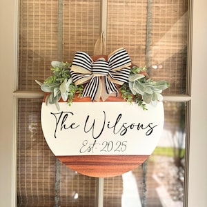 May include: A circular wooden door sign with a white upper section and a brown lower section. The sign displays the text "The Wilsons Est. 2025" in black script. It is adorned with greenery and a black and white striped bow.