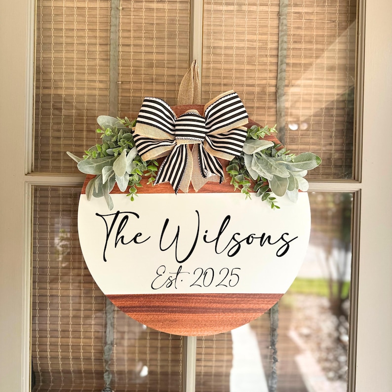 May include: A circular wooden door sign with a white upper section and a brown lower section. The sign displays the text "The Wilsons Est. 2025" in black script. It is adorned with greenery and a black and white striped bow.