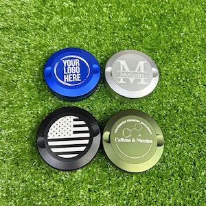 May include: Four round metal objects with different designs. One is blue with "YOUR LOGO HERE", another silver with "M McGuire", a third black with an American flag, and the last green with "Caffeine & Nicotine".