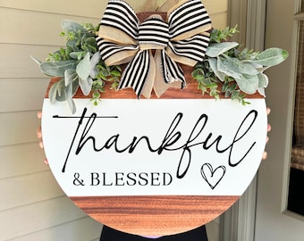 May include: Round wooden sign with the words "Thankful & Blessed" in black script. The sign has a white top section and a brown bottom section. It is decorated with a large bow with black and white stripes and greenery.