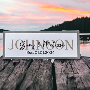 May include: A wooden sign with the text "JOHNSON Gary & Melissa Est. 01.01.2024" in brown lettering on a white background. The sign is sitting on a wooden dock with a lake and forest in the background.