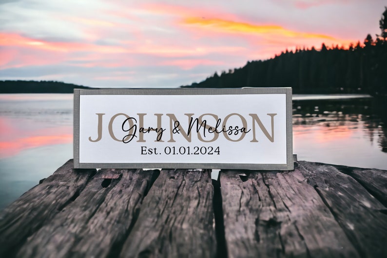May include: A wooden sign with the text "JOHNSON Gary & Melissa Est. 01.01.2024" in brown lettering on a white background. The sign is sitting on a wooden dock with a lake and forest in the background.