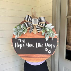 May include: A round wooden door hanger with a black and white striped bow, green foliage, and the text "Hope You Like Dogs" with paw prints.
