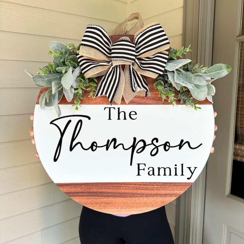 May include: A round wooden door hanger with a white painted center. The center has the text "The Thompson Family" written in black cursive. The top of the hanger is decorated with a black and white striped bow and greenery.