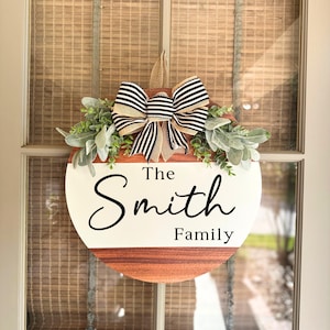 May include: A round wooden door hanger with a white background and the text "The Smith Family" in black cursive script. The hanger is decorated with a black and white striped bow and green foliage.