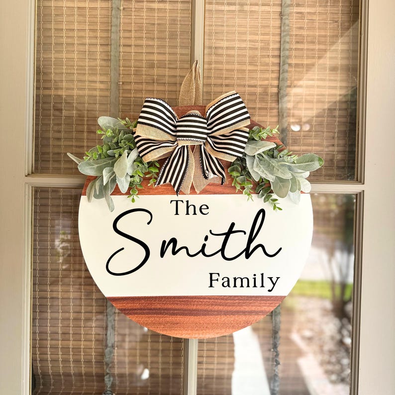 May include: A round wooden door hanger with a white background and the text "The Smith Family" in black cursive script. The hanger is decorated with a black and white striped bow and green foliage.