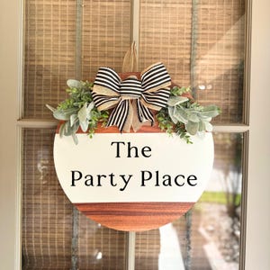 May include: A round wooden door hanger with a white painted center. The center has the text "The Party Place" written in black. The hanger is decorated with a black and white striped bow and greenery.