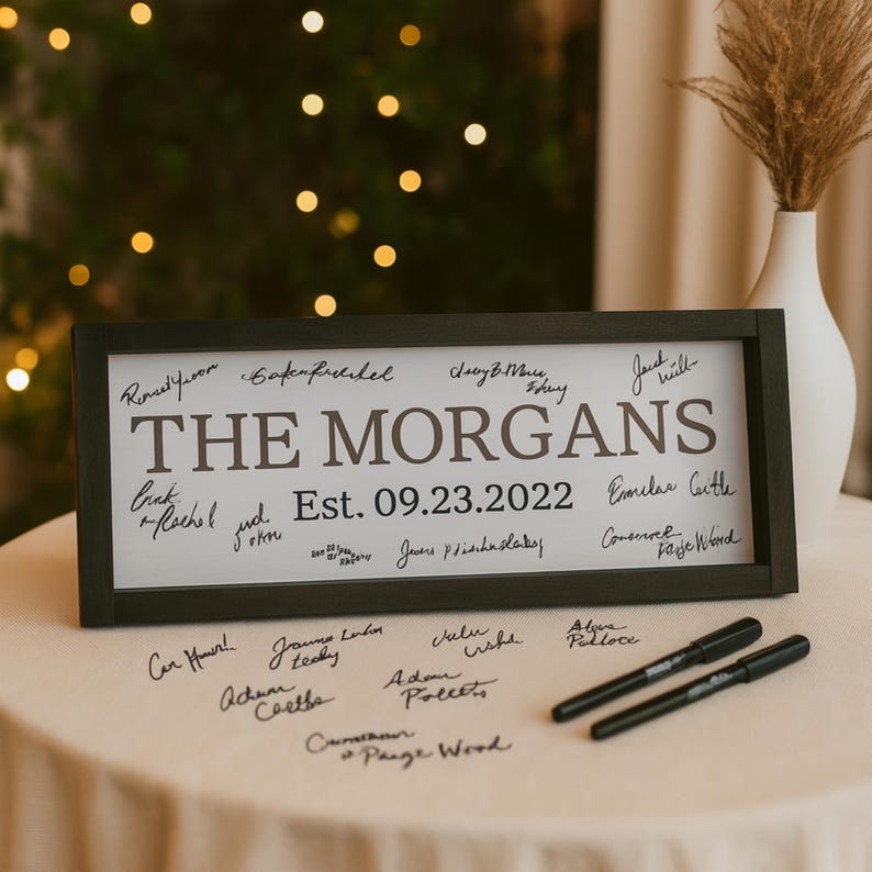 May include: A framed sign with the text "THE MORGANS" and "Est. 09.23.2022" in a black frame. The sign is surrounded by signatures. Two black markers are on the table.