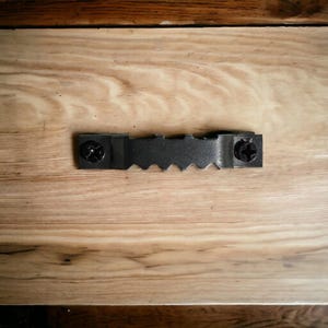 May include: A close-up of a wooden surface with a black metal hanging bracket attached to it. The bracket has two screws and a serrated edge.