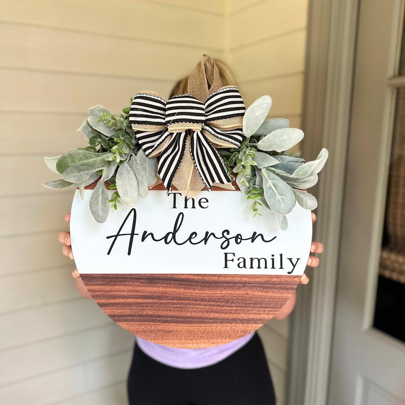 May include: A round wooden door hanger with a white top half and a brown wood bottom half. The top half has the text "The Anderson Family" in black script font. The hanger is decorated with a black and white striped bow and greenery.