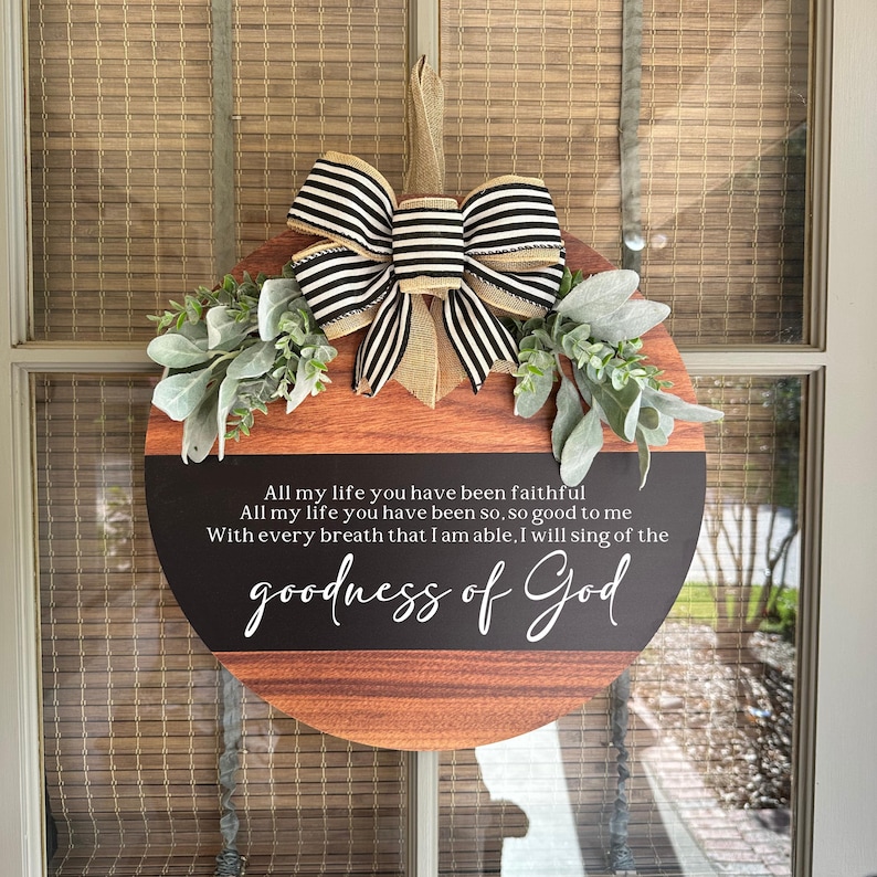 Op de afbeelding: Een rond houten bord met een zwart-wit gestreepte strik en groen. Het bord heeft een zwarte achtergrond met witte tekst die luidt "All my life you have been faithful. All my life you have been so, so good to me. With every breath that I am able. I will sing of the goodness of God."