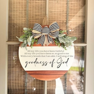 Op de afbeelding: Een rond houten bord met een witte achtergrond en zwarte tekst. Het bord bevat een citaat over de goedheid van God. Het bord is versierd met een zwart-witte strik en groen.