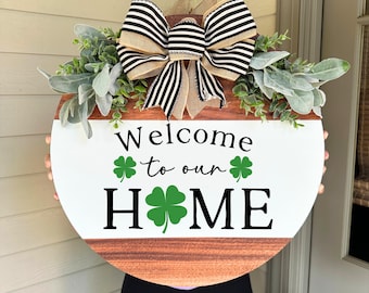 May include: Round wooden welcome sign with the text "Welcome to our HOME" and shamrock accents. The sign features a large bow with black and white stripes and burlap ribbon, and is decorated with greenery.