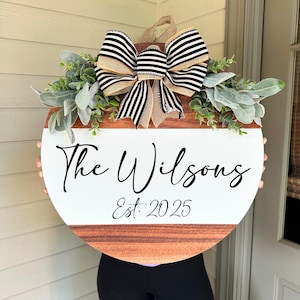 May include: A circular wooden sign with a white central section and a brown wood-grain base. The sign displays the text "The Wilsons" and "Est. 2025" in black script. It is adorned with foliage and a large bow with black and white stripes.
