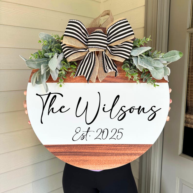 May include: A circular wooden sign with a white central section and a brown wood-grain base. The sign displays the text "The Wilsons" and "Est. 2025" in black script. It is adorned with foliage and a large bow with black and white stripes.
