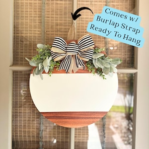 May include: A round wooden sign with a white painted center and a natural wood border. The sign is decorated with a black and white striped bow and greenery. The sign comes with a burlap strap for hanging.