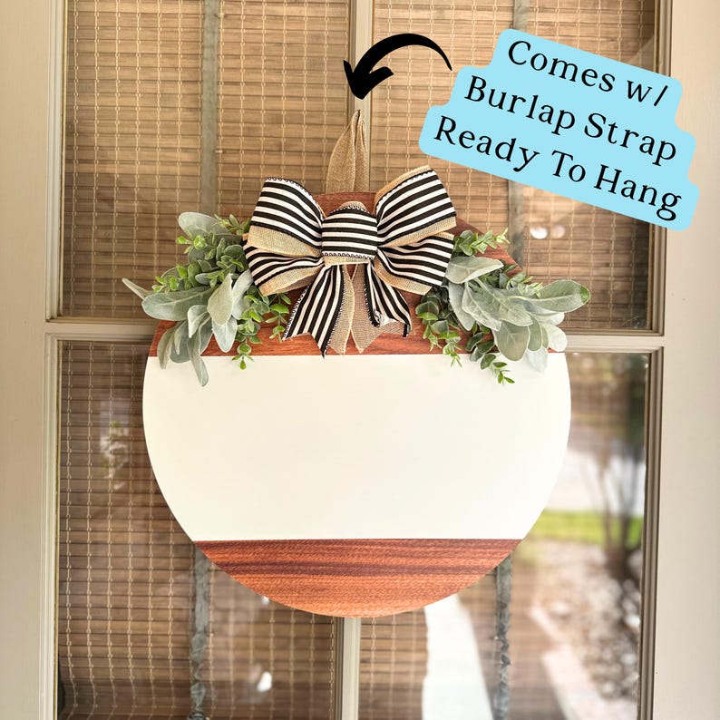 May include: A round wooden sign with a white painted center and a natural wood border. The sign is decorated with a black and white striped bow and greenery. The sign comes with a burlap strap for hanging.