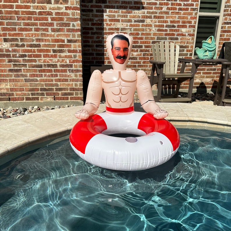 May include: Inflatable pool float shaped like a man with a mustache and a red and white life preserver. The float is in a pool of water, with a brick wall in the background.