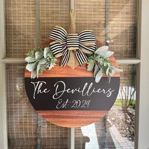 May include: Circular wooden door sign featuring a black and white striped bow, foliage, and the text "The Devilliers Est. 2024". The sign has a wood grain effect and a black stripe with white lettering.