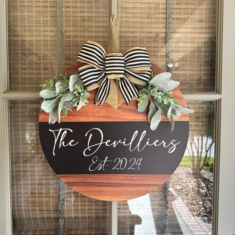 May include: Circular wooden door sign featuring a black and white striped bow, foliage, and the text "The Devilliers Est. 2024". The sign has a wood grain effect and a black stripe with white lettering.
