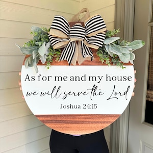 May include: A round wooden sign with the text "As for me and my house we will serve the Lord Joshua 24:15". The sign has a white top and a brown wood-grain bottom. It is decorated with greenery and a large bow with black and white stripes.
