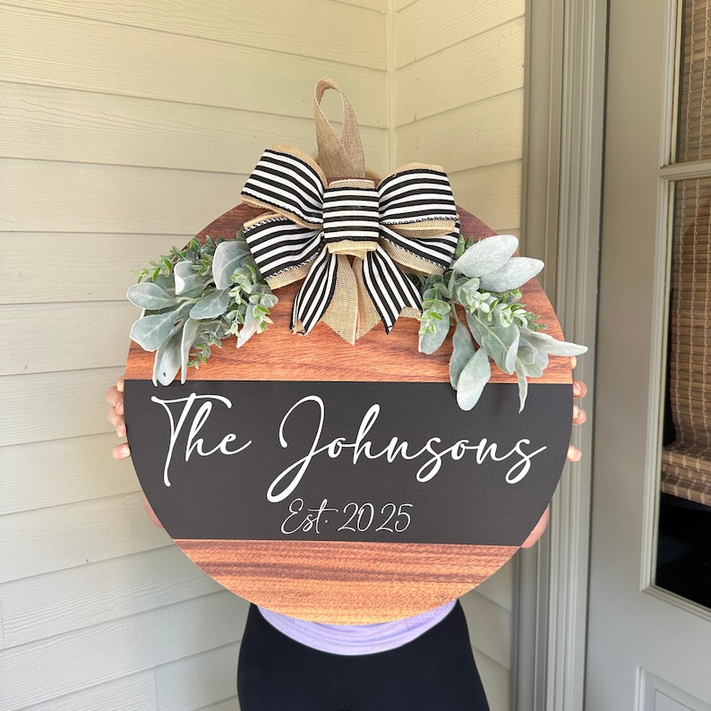 May include: A circular wooden sign featuring a black and white striped bow and decorative greenery. The sign displays the name "The Johnsons" and the year "Est. 2025" in white script on a black banner. The sign has a brown wood grain finish.