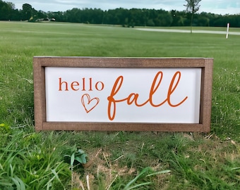 May include: A wooden sign with the text "hello fall" in orange lettering with a heart symbol. The sign is sitting on green grass.