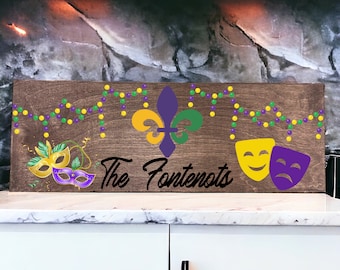 Puede incluir: Un letrero de madera con el texto "The Fontenots" en letras cursivas negras. El letrero está decorado con cuentas de Mardi Gras moradas, verdes y doradas, una flor de lis y dos máscaras.