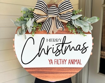 Può includere: Un cartello natalizio rotondo in legno con la scritta "MERRY Christmas YA FILTHY ANIMAL". Il cartello presenta un design bianco e marrone, un fiocco a righe e verde.