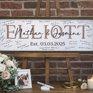 May include: A wooden sign with the names "Nathan & Jasmine" and the date "03.03.2025" displayed on an easel. The sign is surrounded by a bouquet of flowers, a framed photo, a pen, rings, and a candle.