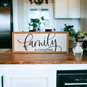 May include: A rectangular wooden sign with the words "Family is Everything" in black script. The sign has a white background and a light brown wooden frame. The sign is displayed on a wooden countertop.