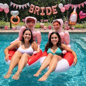 May include: Two women in a pool, sitting on red and white inflatable rings. Two people in pink inflatable costumes stand behind them. The word "BRIDE" is spelled out in large gold balloons, with other pink balloons and decorations.
