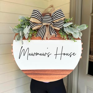 May include: A round wooden door hanger with a white background and the text "Mawmaw's House" in black script. The door hanger is decorated with a black and white striped bow and greenery.