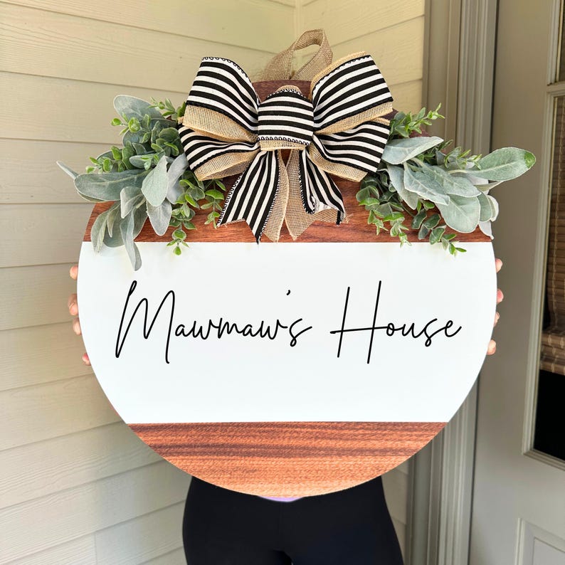 May include: A round wooden door hanger with a white background and the text "Mawmaw's House" in black script. The door hanger is decorated with a black and white striped bow and greenery.