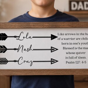 May include: A rectangular wooden sign with a white background featuring three black arrow graphics. Each arrow is labeled with the names Lola, Nash, and Cruz. The text reads: Like arrows in the hands of a warrior are children born in one's youth. Blessed is the man whose quiver is full of them. Psalm 127 : 4-5.