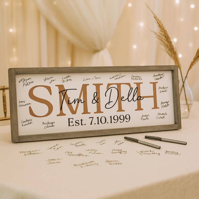 May include: A rectangular wooden sign with the names "Smith & Della" in brown and black lettering, with the date "Est. 7.10.1999" below. The sign is framed in gray and has signatures. Two black pens are on the table.