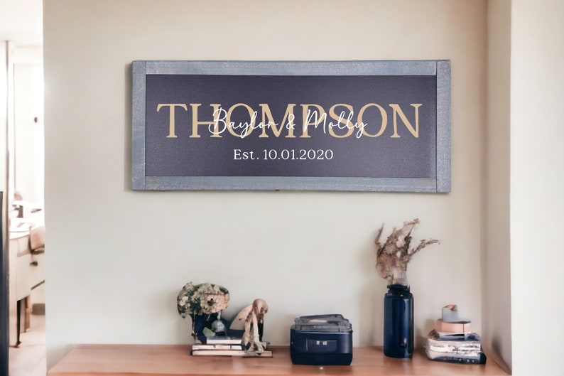 May include: A framed sign with the last name "Thompson" in gold lettering, followed by "Baylon & Molly" in white cursive lettering and "Est. 10.01.2020" in gold lettering.