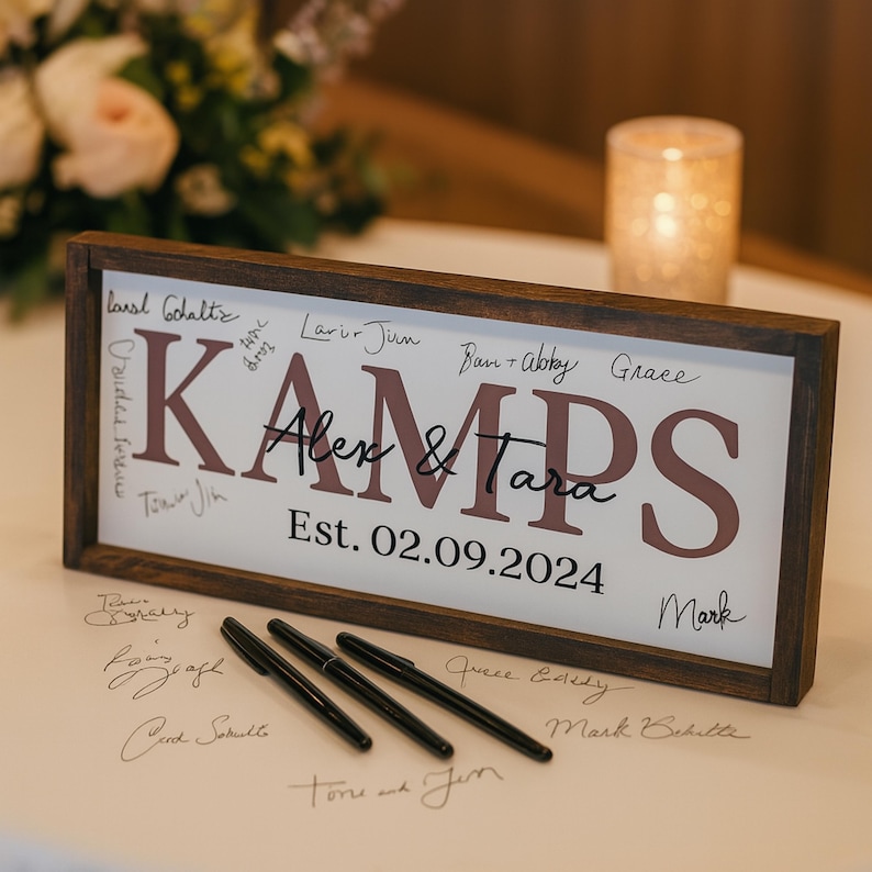 May include: A wooden framed sign with the name "KAMPS" in mauve letters, with "Alex & Tara" and the date "Est. 02.09.2024" in black script. Several signatures and three black pens are visible.