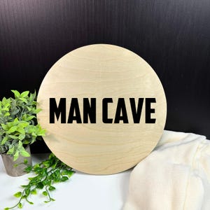 May include: A round wooden sign with the text "MAN CAVE" in black lettering.