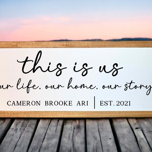 May include: A white wooden sign with black text that reads "this is us our life, our home, our story CAMERON BROOKE ARI EST. 2021". The sign is sitting on a wooden surface.