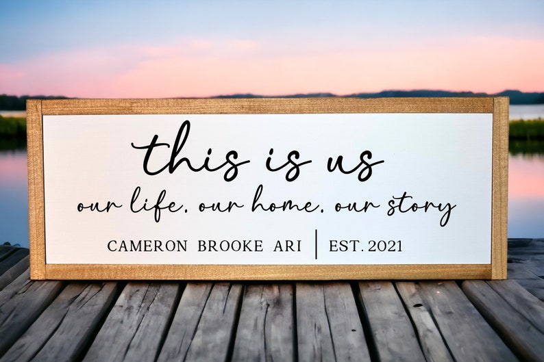 May include: A white wooden sign with black text that reads "this is us our life, our home, our story CAMERON BROOKE ARI EST. 2021". The sign is sitting on a wooden surface.