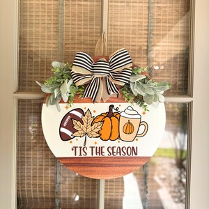 May include: Round wooden door hanger with a cream-colored top and a brown bottom. The design includes a football, pumpkins, a leaf, and a coffee cup. A black and white striped bow and greenery are at the top. The text reads "'Tis the Season."