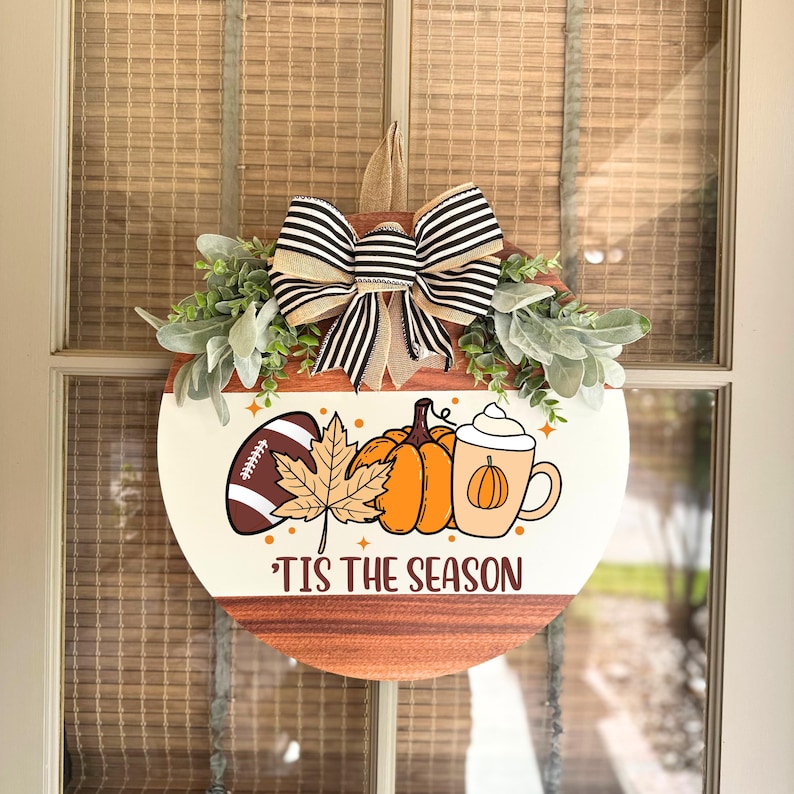 May include: Round wooden door hanger with a cream-colored top and a brown bottom. The design includes a football, pumpkins, a leaf, and a coffee cup. A black and white striped bow and greenery are at the top. The text reads "'Tis the Season."
