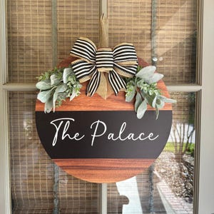 May include: A round wooden door hanger with a black and white striped bow and greenery. The door hanger has a black chalkboard section with the text "The Palace" written in white.