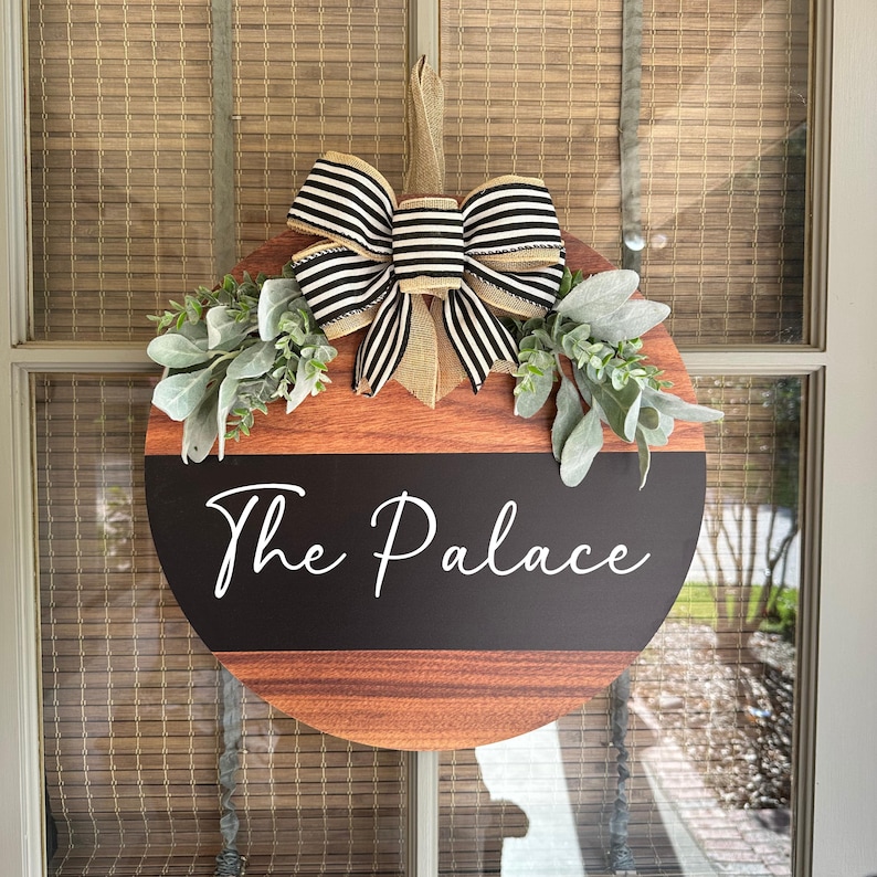 May include: A round wooden door hanger with a black and white striped bow and greenery. The door hanger has a black chalkboard section with the text "The Palace" written in white.